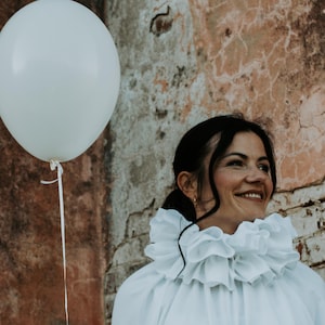 May include: A person wearing a white ruffled collar shirt with a large black pom-pom button. A white balloon floats in the air next to the person. The background is a weathered brick wall.