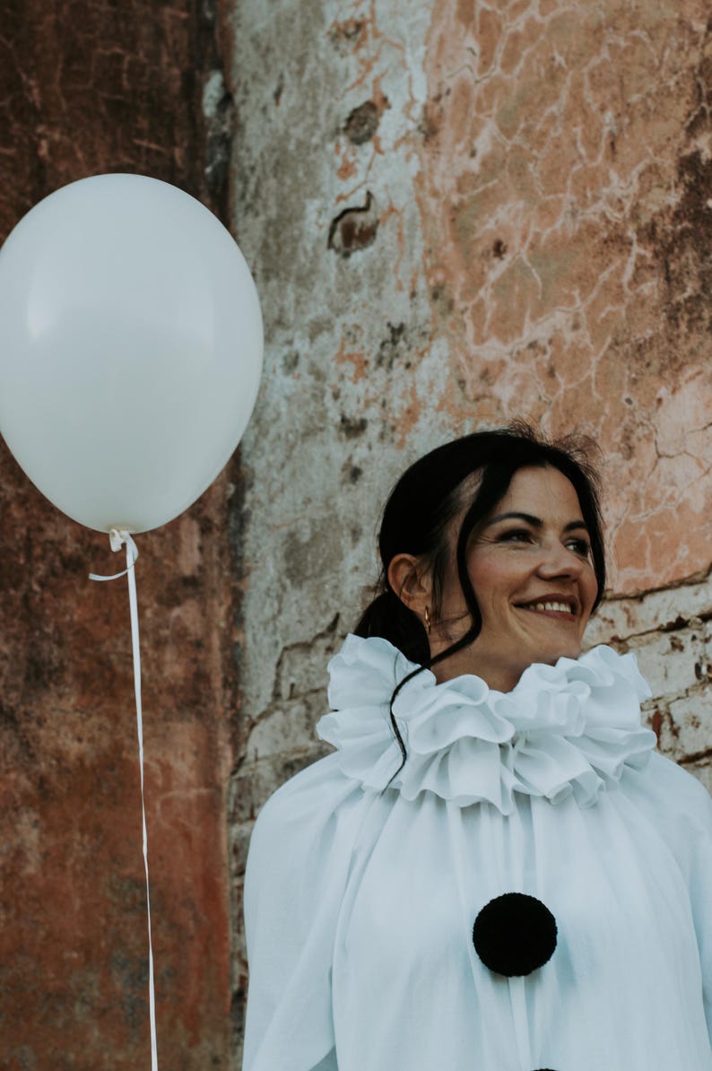May include: A person wearing a white ruffled collar shirt with a large black pom-pom button. A white balloon floats in the air next to the person. The background is a weathered brick wall.