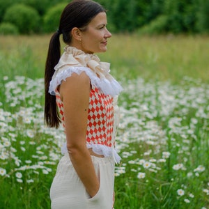 May include: A woman wearing a white and red checkered top with a white ruffled collar and white pants. She is standing in a field of white daisies.