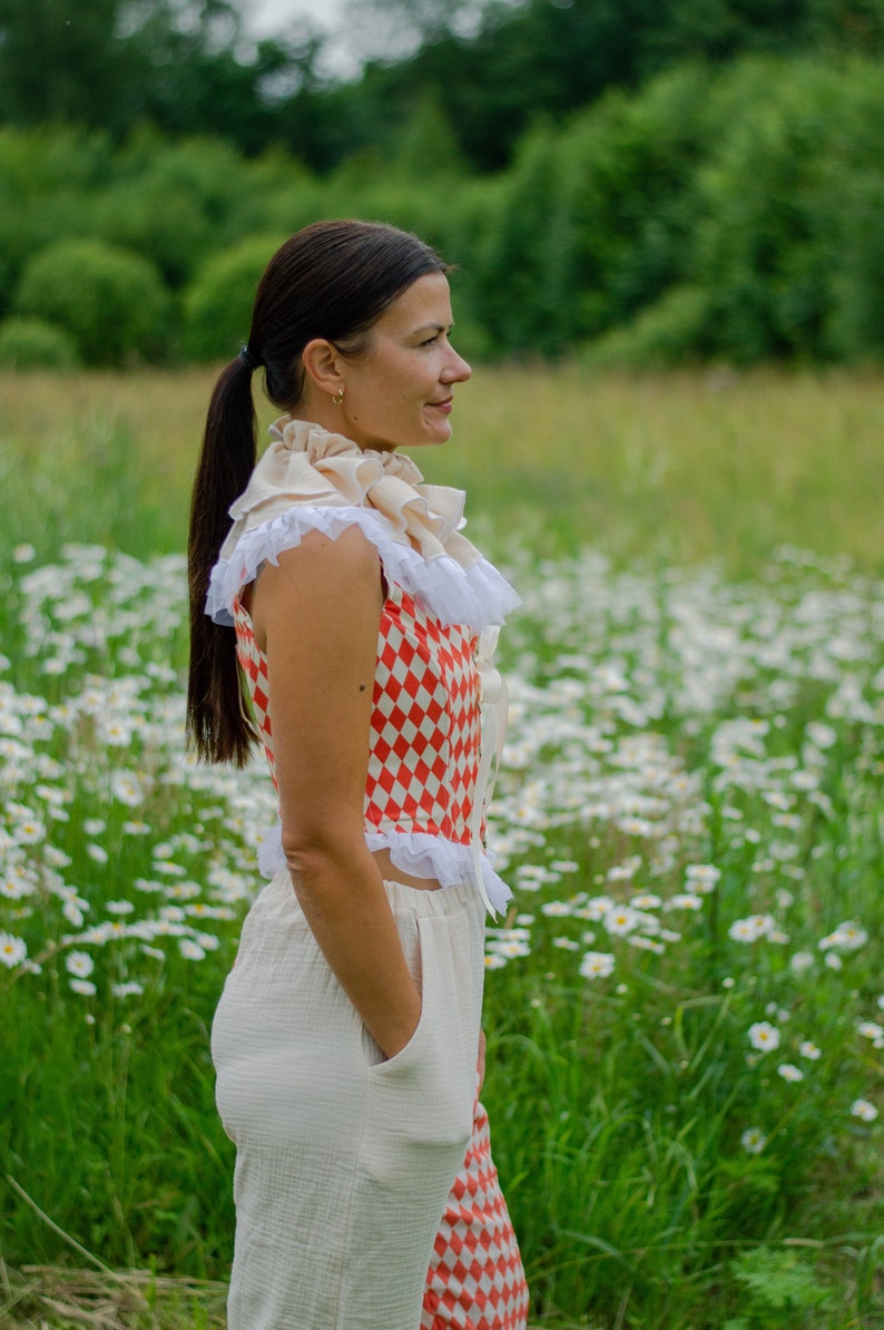 May include: A woman wearing a white and red checkered top with a white ruffled collar and white pants. She is standing in a field of white daisies.