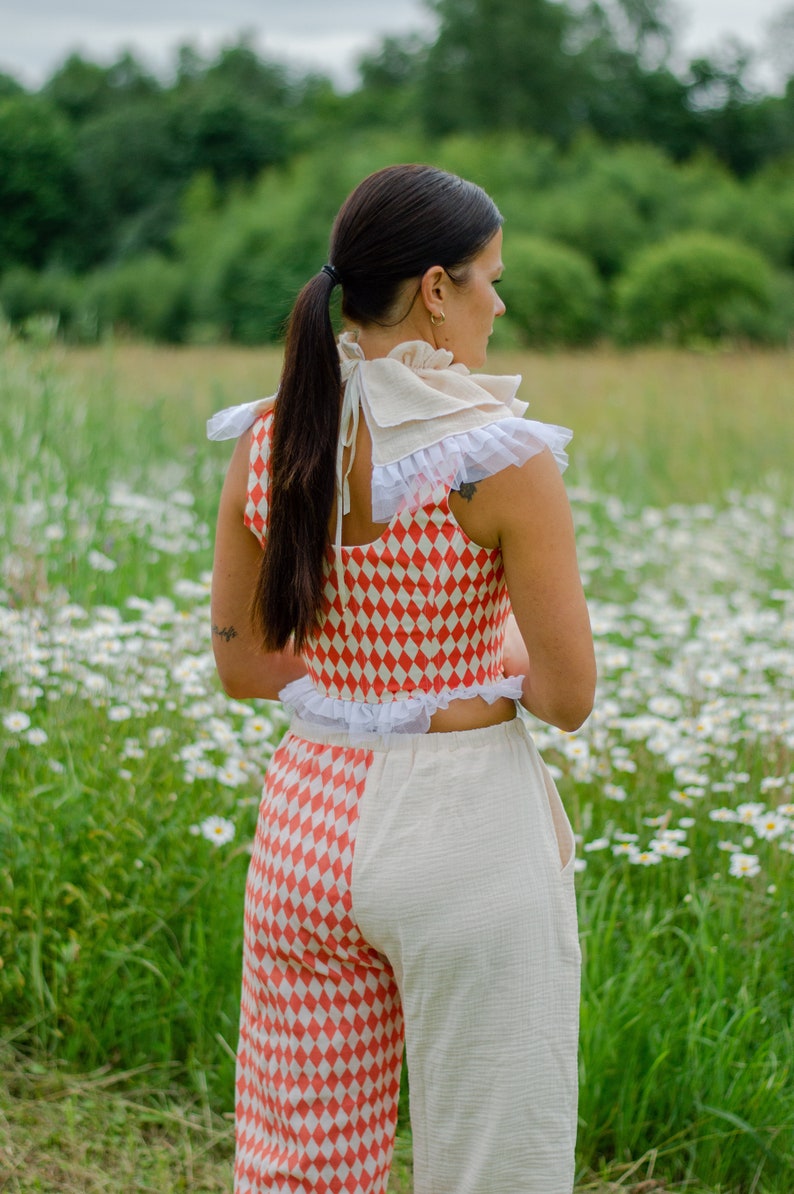 May include: A woman wearing a red and white checkered top with a white lace trim and matching pants. The top is a cropped style and the pants are high-waisted. She is wearing a white lace collar with a bow in the back. The woman is standing in a field of daisies.