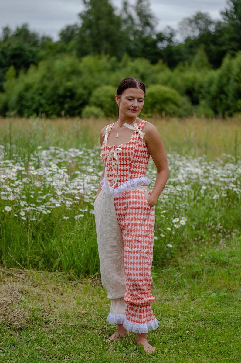 May include: A woman wearing a red and white checkered top with a white ruffle and matching pants. The pants are split, with one leg being white and the other red and white checkered. She is standing in a field of white flowers.