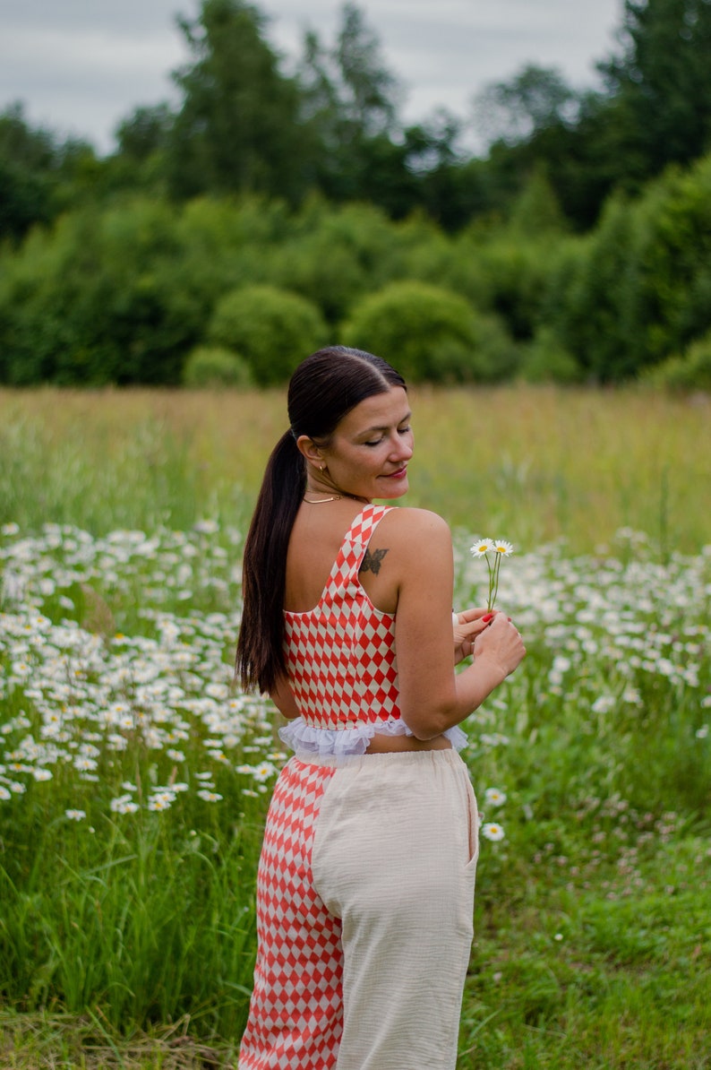 May include: A woman wearing a red and white checkered crop top with a white ruffle trim and matching pants. She is holding a small bouquet of white daisies in her right hand. The woman is standing in a field of daisies.