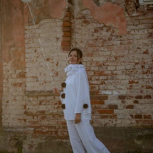 May include: A person in a white clown costume stands in front of a weathered brick wall, holding a white balloon. The costume includes a ruffled collar and large black buttons. The setting is outdoors with green grass.