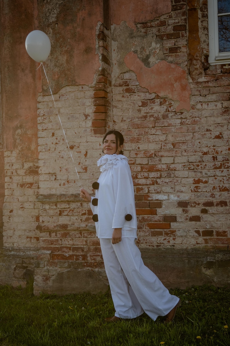 May include: A person in a white clown costume stands in front of a weathered brick wall, holding a white balloon. The costume includes a ruffled collar and large black buttons. The setting is outdoors with green grass.