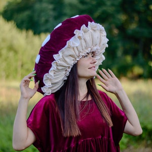 May include: A woman wearing a burgundy velvet dress with a large mushroom-shaped hat. The hat is made of burgundy velvet and white fabric. The woman is looking up and smiling.