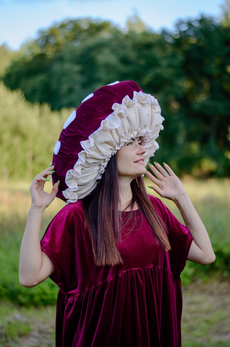 May include: A woman wearing a burgundy velvet dress with a large mushroom-shaped hat. The hat is made of burgundy velvet and white fabric. The woman is looking up and smiling.