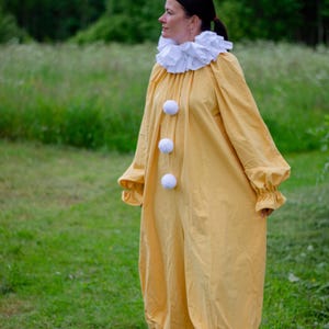 May include: A yellow clown costume with a white ruffled collar and three white pom-poms down the front. The long-sleeved jumpsuit has gathered cuffs and ankles. The person is wearing brown shoes and standing in a grassy field.