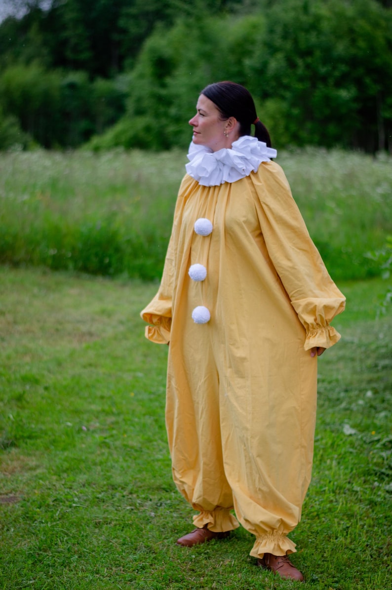May include: A yellow clown costume with a white ruffled collar and three white pom-poms down the front. The long-sleeved jumpsuit has gathered cuffs and ankles. The person is wearing brown shoes and standing in a grassy field.