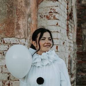 May include: A person in a white clown costume with a ruffled collar and two large black pom-poms. They are holding a white balloon. The background is a weathered brick wall.
