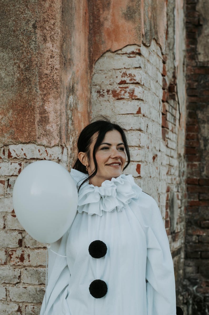 May include: A person in a white clown costume with a ruffled collar and two large black pom-poms. They are holding a white balloon. The background is a weathered brick wall.