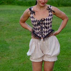 May include: A beige and black diamond-patterned corset top featuring black ribbon shoulder ties, a front lace-up closure, and a black tulle hem trim, paired with solid beige high-waisted bloomers with gathered leg openings and a ruffled edge.