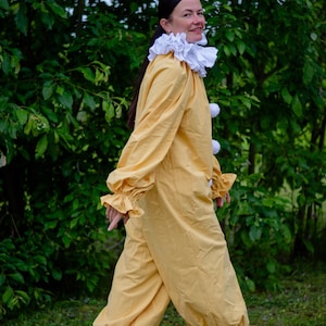 May include: A yellow clown costume with a white ruffled collar and white pom-poms. The long-sleeved jumpsuit has ruffled cuffs and ankles. The person is wearing brown shoes and walking on grass.