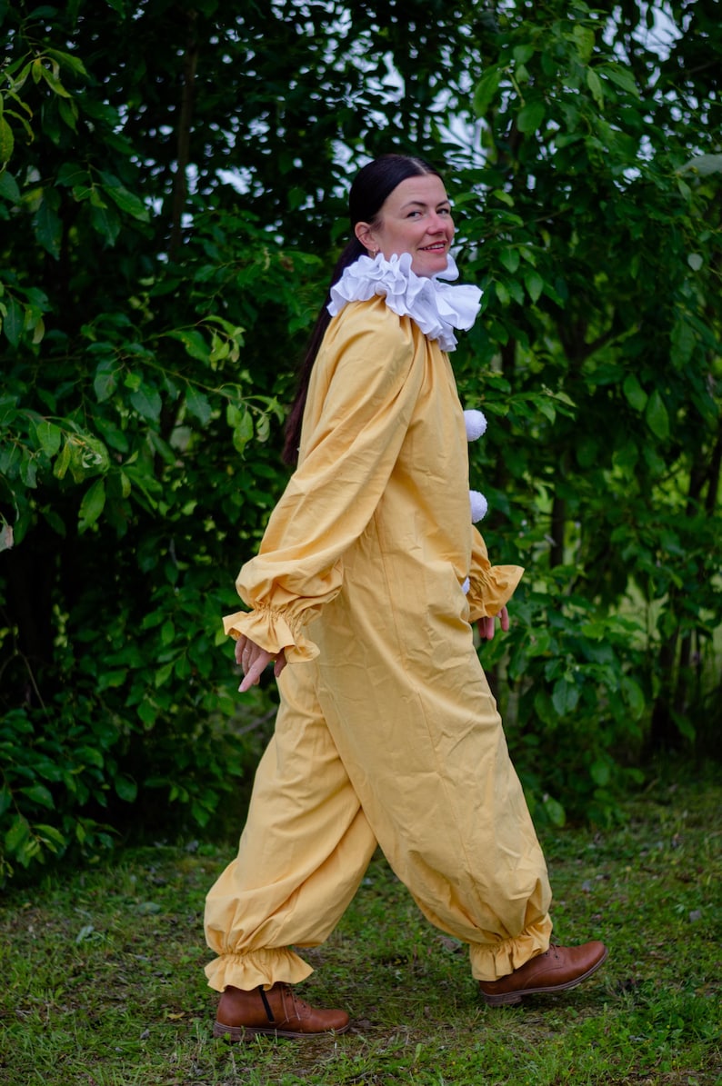 May include: A yellow clown costume with a white ruffled collar and white pom-poms. The long-sleeved jumpsuit has ruffled cuffs and ankles. The person is wearing brown shoes and walking on grass.