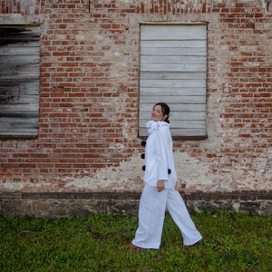 May include: A person wearing a white outfit with a ruffled collar and black buttons, walking in front of a weathered brick wall. The outfit includes a long-sleeved jacket and matching pants. The background features boarded-up windows and green grass.