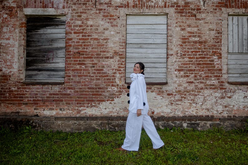 May include: A person wearing a white outfit with a ruffled collar and black buttons, walking in front of a weathered brick wall. The outfit includes a long-sleeved jacket and matching pants. The background features boarded-up windows and green grass.