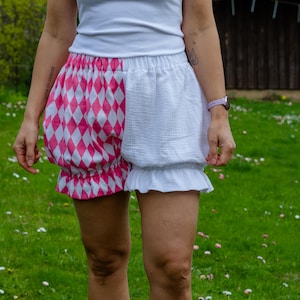 May include: High-waisted bloomers featuring a split design with one side in a pink and white diamond harlequin pattern and the other side in solid white. The garment has an elasticized waistband and ruffled leg openings with a textured fabric.