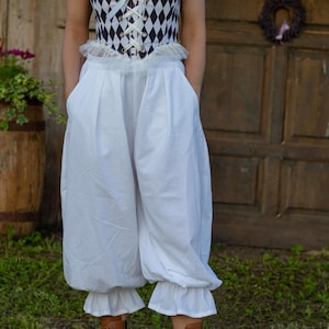 May include: A white and black corset-style top with a diamond pattern is paired with white, puffy pants. The pants have a ruffled hem. The outfit is part of a costume or historical clothing ensemble.