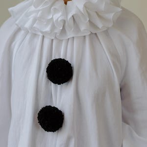 May include: White clown shirt with a ruffled collar and three large black pom-pom buttons. The shirt is made of a soft, flowing fabric, perfect for a costume or theatrical performance.