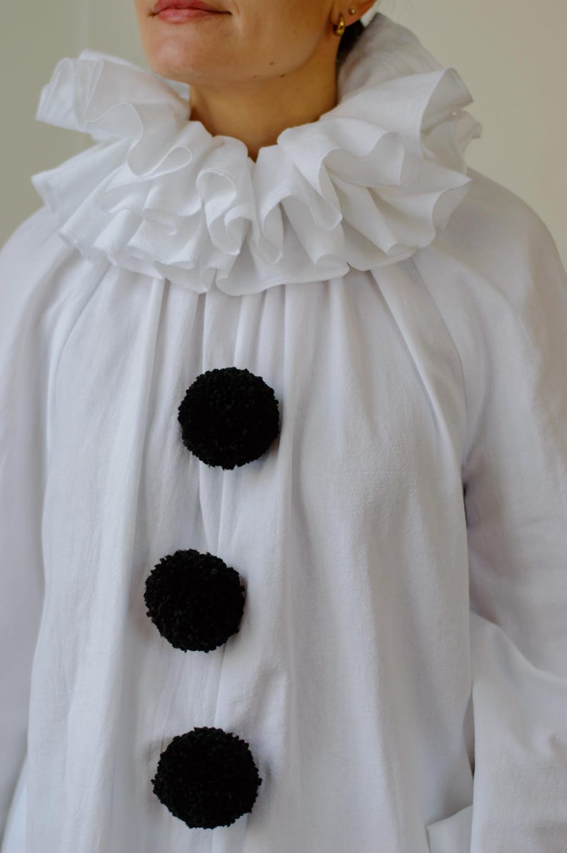 May include: White clown shirt with a ruffled collar and three large black pom-pom buttons. The shirt is made of a soft, flowing fabric, perfect for a costume or theatrical performance.