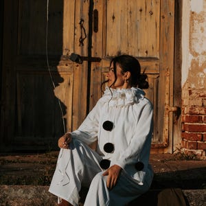 May include: A person in a white clown costume with a ruffled collar and large black buttons sits on stone steps, holding a white balloon. The background features a weathered wooden door and brick wall, with warm sunlight.