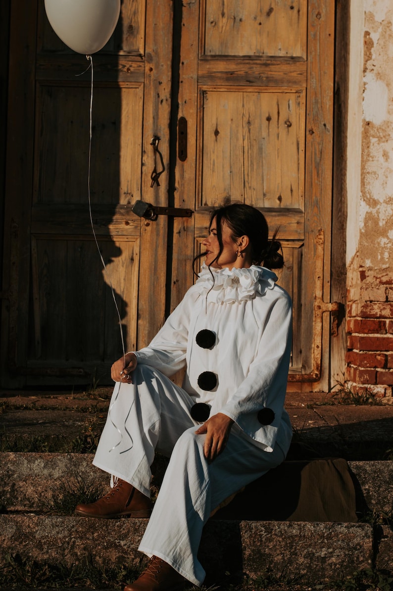 May include: A person in a white clown costume with a ruffled collar and large black buttons sits on stone steps, holding a white balloon. The background features a weathered wooden door and brick wall, with warm sunlight.