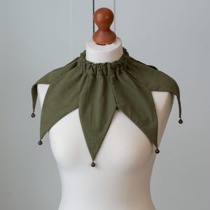 Op de afbeelding: Olijfgroene narrenkraag met puntige randen en kleine bruine kralen aan de uiteinden. De kraag is bij de halslijn gerimpeld en wordt getoond op een witte mannequin. Bovenop de mannequin staat een houten hoofd.