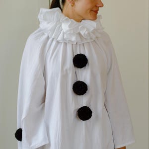 May include: White clown costume with a ruffled collar and large black pom-pom buttons. The long-sleeved shirt has a relaxed fit. The costume is suitable for Halloween or costume parties.