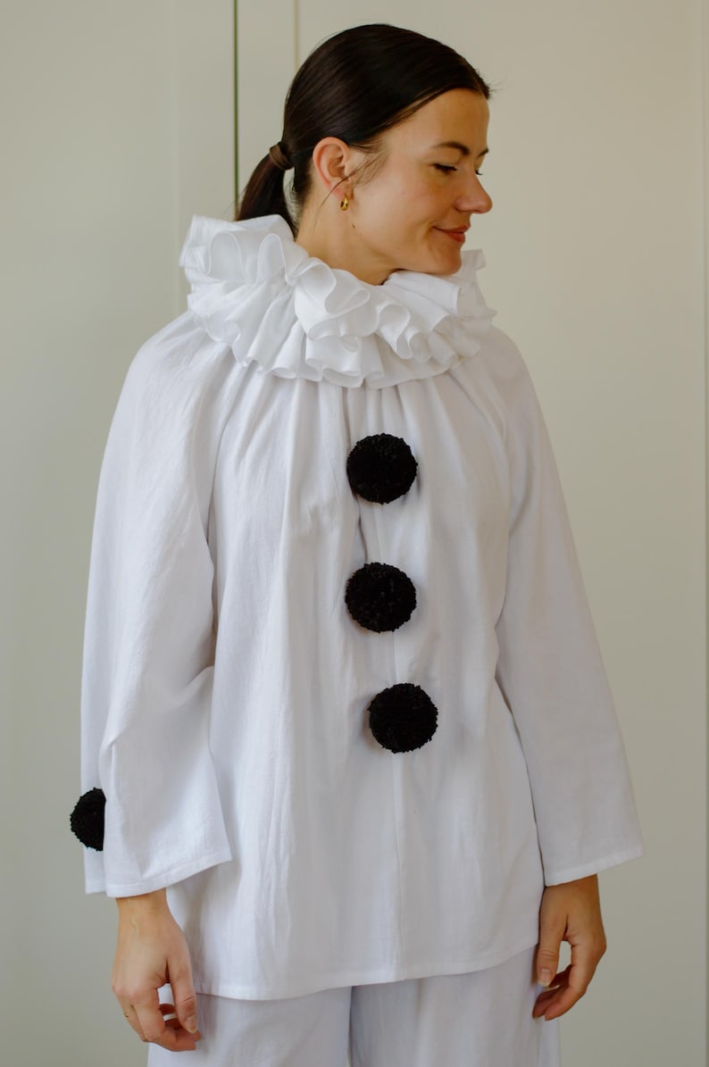 May include: White clown costume with a ruffled collar and large black pom-pom buttons. The long-sleeved shirt has a relaxed fit. The costume is suitable for Halloween or costume parties.