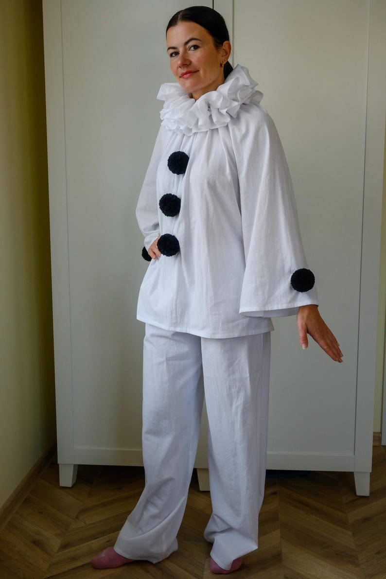 May include: White clown costume featuring a ruffled collar, large black pom-poms, and wide-leg pants. The costume includes a long-sleeved top and is suitable for theatrical performances or costume parties.