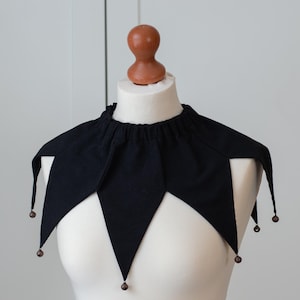 May include: A black jester collar with a series of downward-pointing, diamond-shaped points. Each point has a small, round, brown bead at the tip. The collar is displayed on a white mannequin head and shoulders.