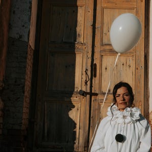 May include: A person in a white clown costume with a ruffled collar and large black buttons, holding a white balloon. The background features a weathered wooden door and a brick wall, with a shadow on the door.