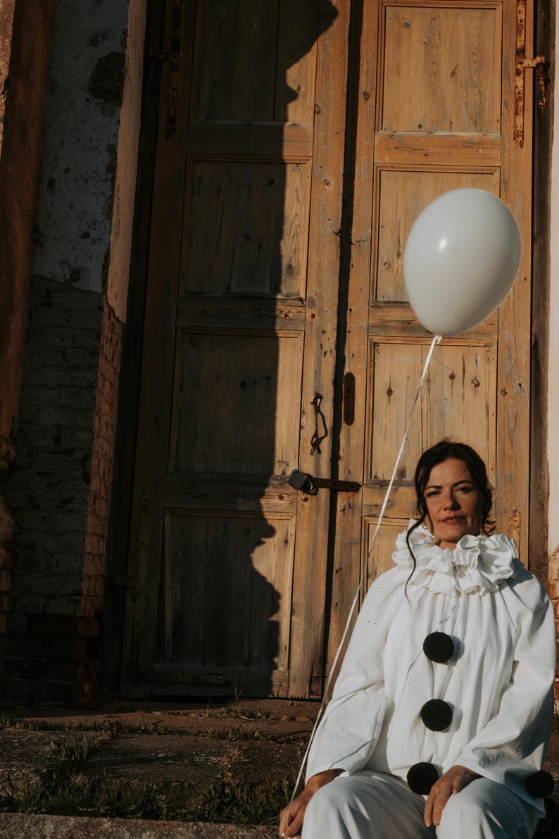 May include: A person in a white clown costume with a ruffled collar and large black buttons, holding a white balloon. The background features a weathered wooden door and a brick wall, with a shadow on the door.