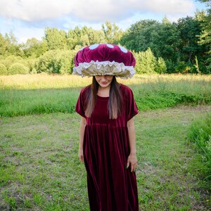 May include: A woman wearing a burgundy velvet dress and a large, red and white mushroom-shaped hat stands in a grassy field.