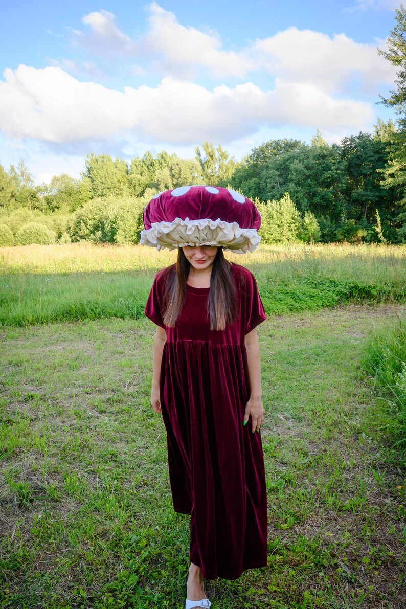 May include: A woman wearing a burgundy velvet dress and a large, red and white mushroom-shaped hat stands in a grassy field.