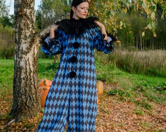 May include: A blue and black diamond-patterned clown jumpsuit with a black ruffled collar and large black pom-pom buttons. The costume has long sleeves and is worn with black shoes. The background includes a tree, pumpkins, and autumn foliage.