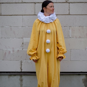 May include: A yellow clown costume with a white ruffled collar and three white pom-poms down the front. The long-sleeved jumpsuit has ruffled cuffs and ankles. The person is wearing brown shoes.