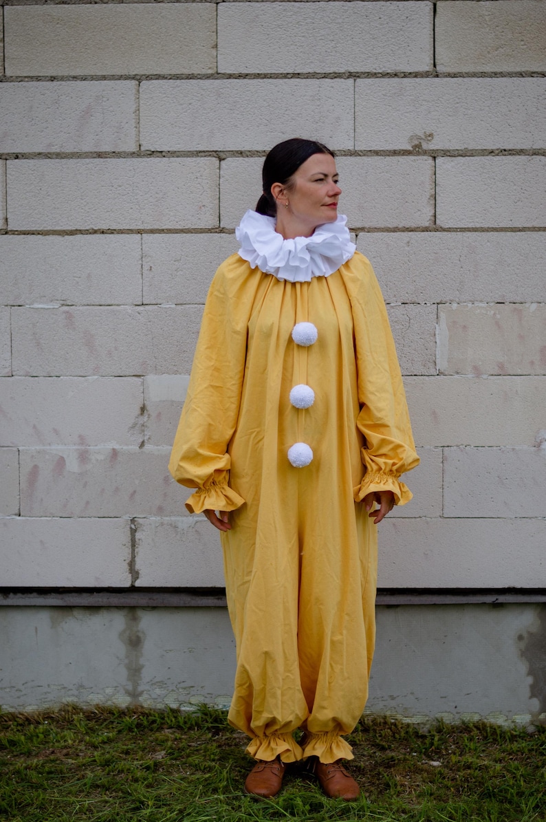 May include: A yellow clown costume with a white ruffled collar and three white pom-poms down the front. The long-sleeved jumpsuit has ruffled cuffs and ankles. The person is wearing brown shoes.