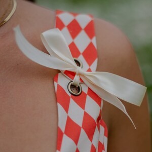 May include: A red and white checkered fabric with a white satin ribbon tied in a bow. The ribbon is tied around two metal eyelets.