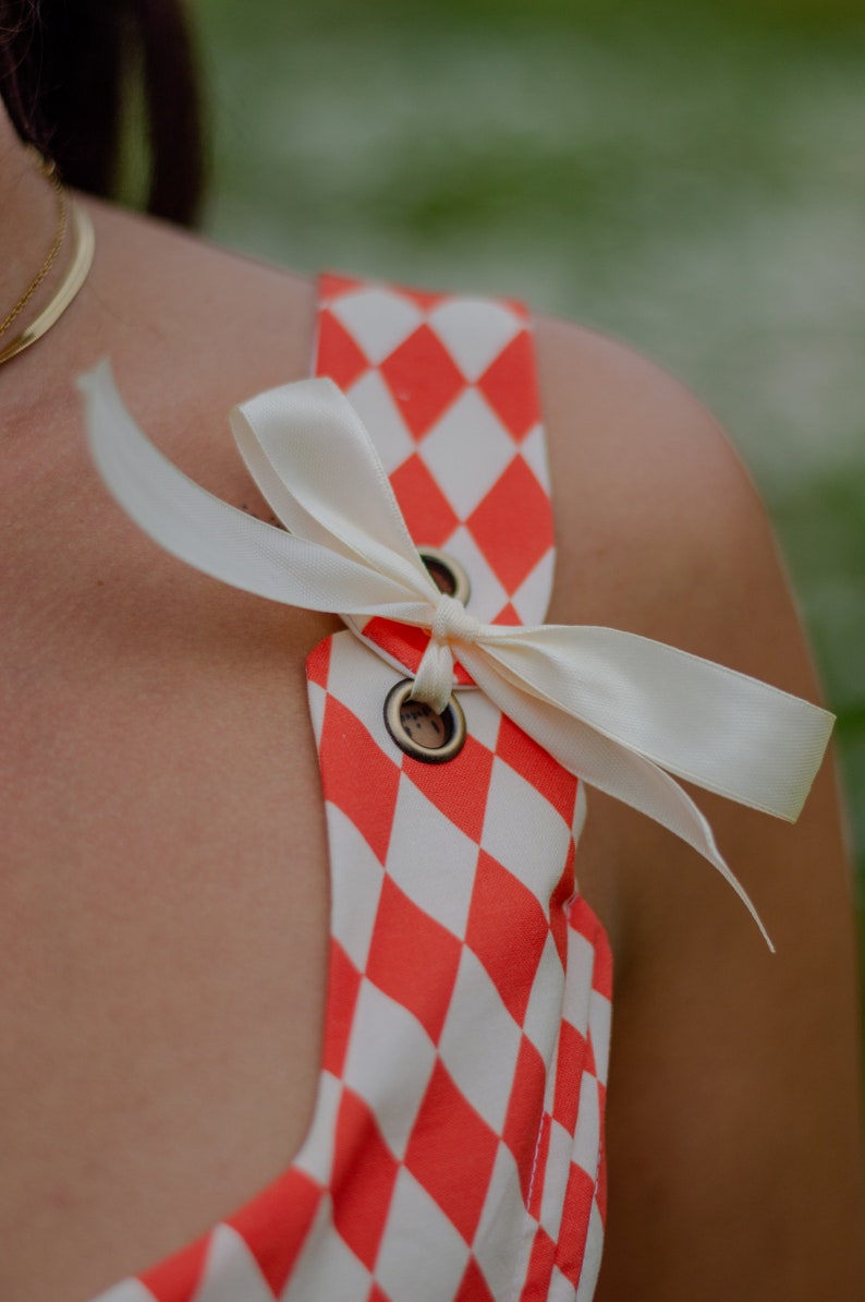 May include: A red and white checkered fabric with a white satin ribbon tied in a bow. The ribbon is tied around two metal eyelets.