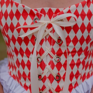 May include: A red and white checkered corset with a white satin ribbon tied in a bow at the front. The corset has a lace-up front with metal eyelets.