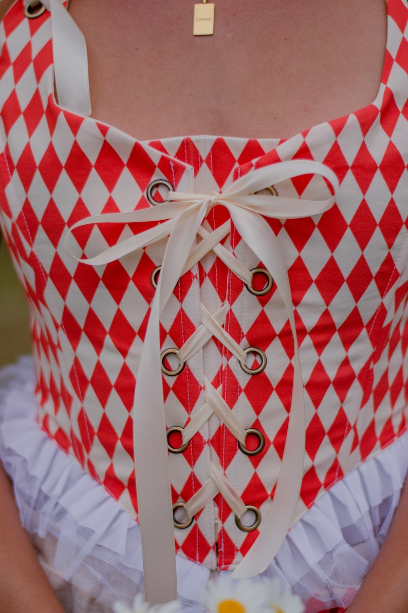May include: A red and white checkered corset with a white satin ribbon tied in a bow at the front. The corset has a lace-up front with metal eyelets.