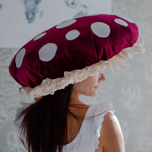 May include: A woman wearing a large, red velvet mushroom hat with white polka dots and a cream-colored ruffle trim.
