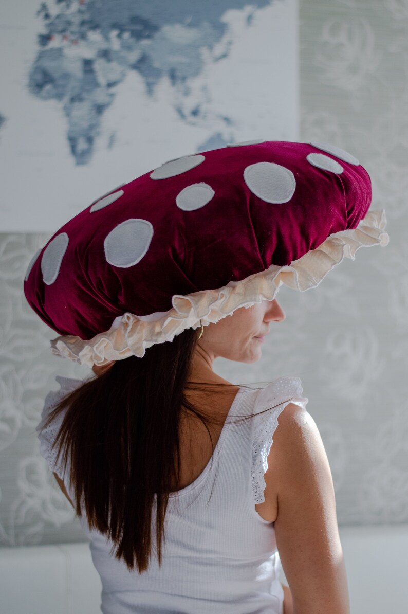 May include: A woman wearing a large, red velvet mushroom hat with white polka dots and a cream-colored ruffle trim.