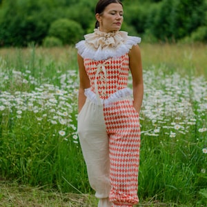 May include: A woman wearing a red and white checkered jumpsuit with a white ruffled collar. The jumpsuit has a lace trim and is cinched at the waist with a ribbon. The woman is standing in a field of daisies.