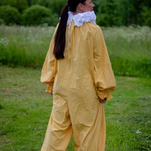 May include: A yellow jumpsuit with long sleeves and ruffled cuffs and ankles. The costume has a white ruffled collar. The person is standing in a grassy field with trees in the background. The outfit is reminiscent of a Renaissance or theatrical costume.