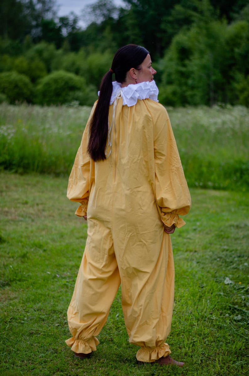May include: A yellow jumpsuit with long sleeves and ruffled cuffs and ankles. The costume has a white ruffled collar. The person is standing in a grassy field with trees in the background. The outfit is reminiscent of a Renaissance or theatrical costume.