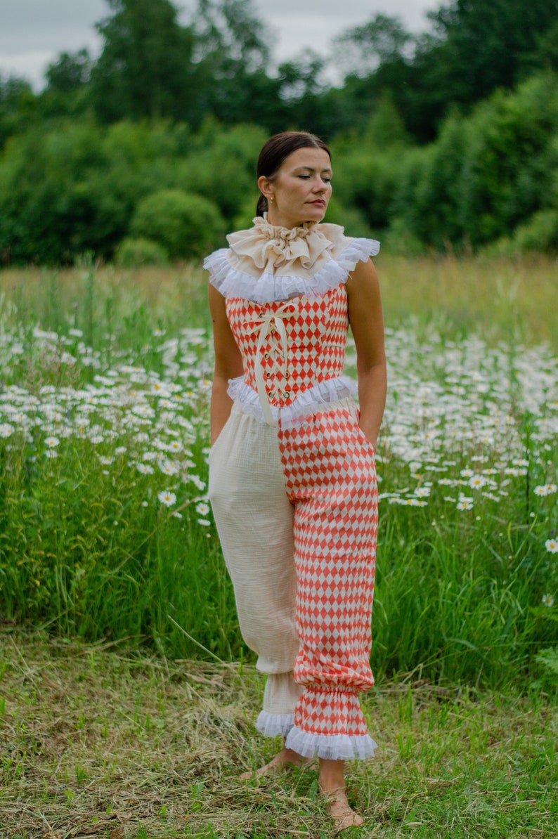 May include: A woman wearing a red and white checkered jumpsuit with a white ruffled collar and a white lace trim. She is standing in a field of daisies.
