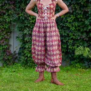 May include: A two-piece outfit consisting of a sleeveless corset-style top with white lace-up front detailing and matching loose-fitting, high-waisted trousers featuring a repeating diamond pattern in shades of red, gold, and dark brown.
