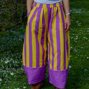 May include: Wide-leg trousers featuring vertical yellow and purple stripes with a solid purple hem at the bottom. The pants have an elastic waistband and side pockets, paired with a white sleeveless top featuring a purple collar detail.